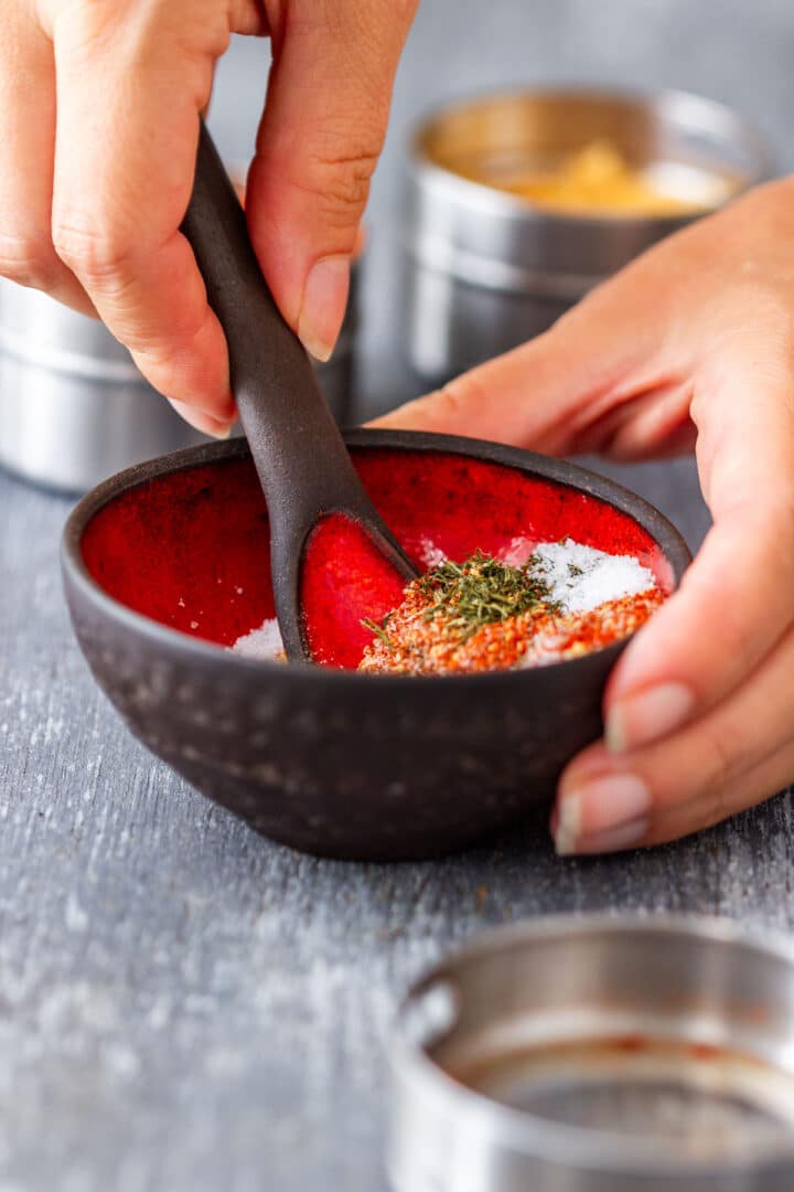 Hand stirring spices together with a small spoon in a black bowl.