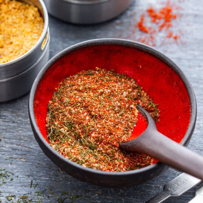 Homemade fish seasoning in a black bowl with a measuring spoon and dried dill scattered nearby.