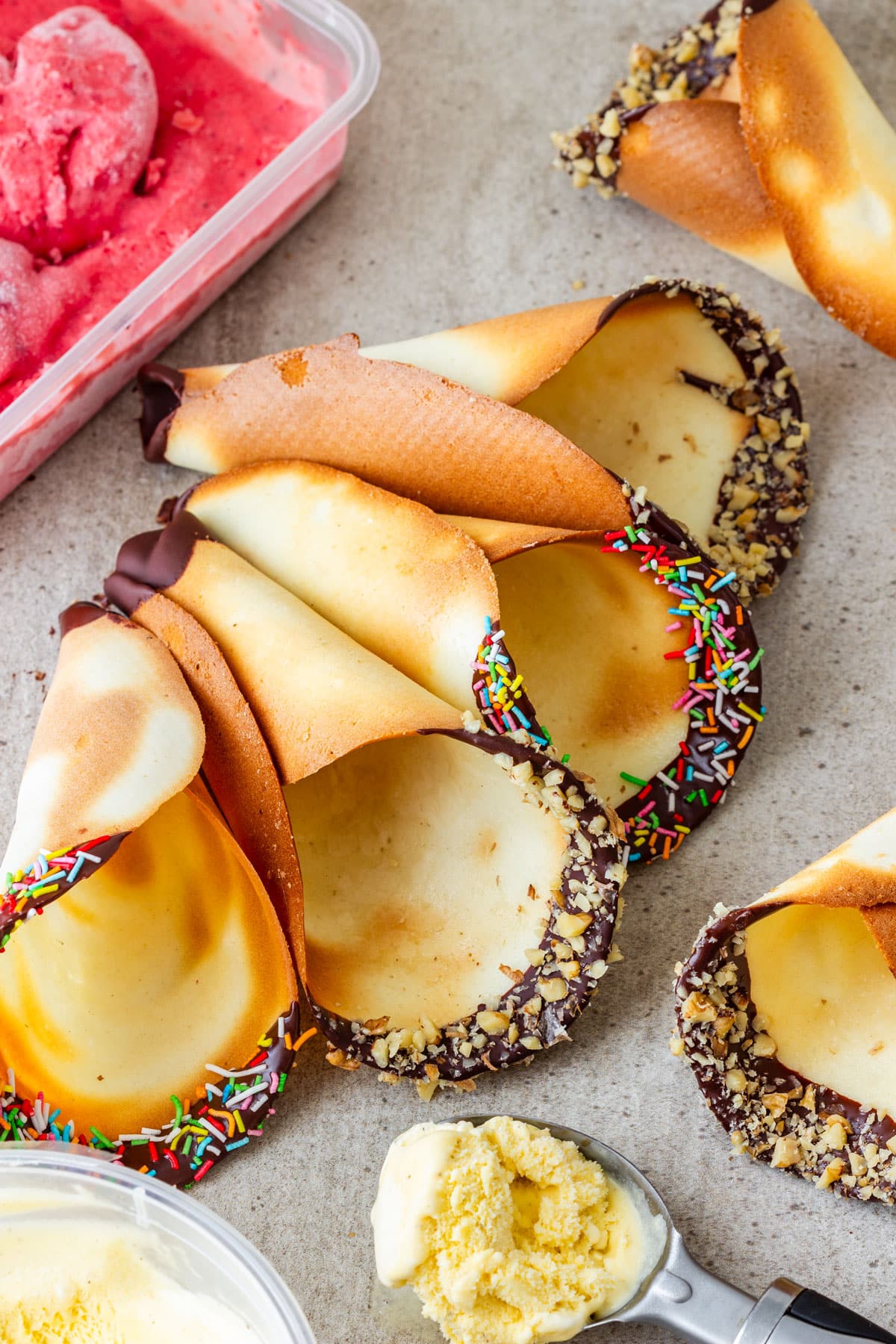 Homemade ice cream cones with chocolate-dipped rims, walnuts and sprinkles, next to strawberry and vanilla ice cream.