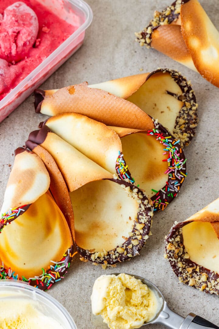 Learn how to make homemade ice cream cones in the oven without a waffle maker. These thin, crisp cones have chocolate-dipped rims with chopped nuts or colorful sprinkles, plus a marshmallow tucked into the tip to help prevent leaks. Fill them with strawberry, vanilla or any ice cream you like.