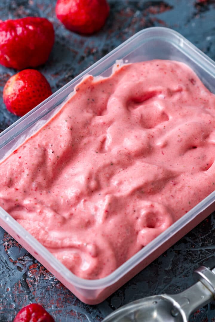 Soft strawberry ice cream in a freezer container with frozen strawberries nearby.