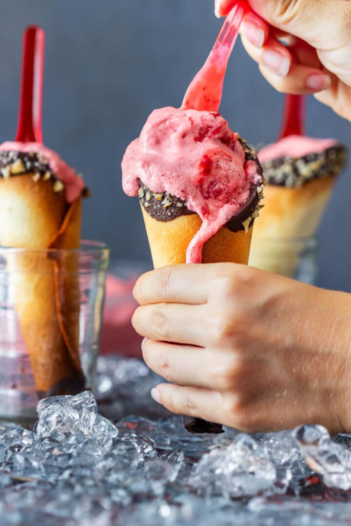 A spoon lifting melting strawberry ice cream from a chocolate-dipped cone.