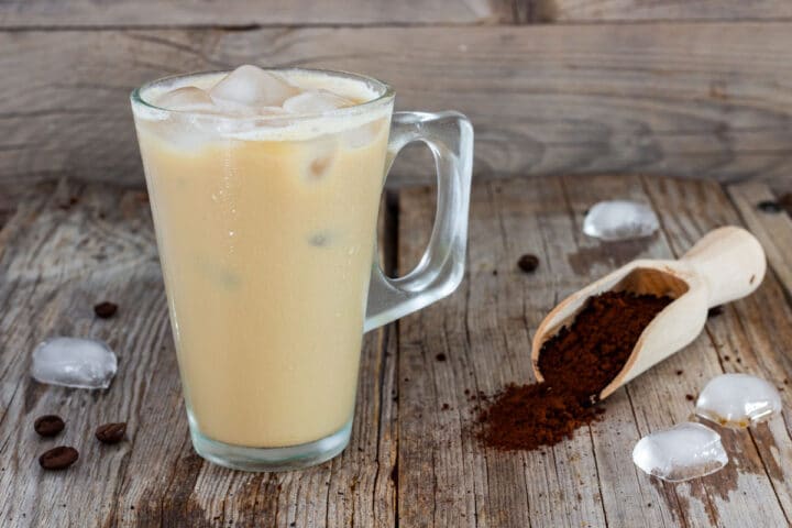Glass of iced coffee beside a wooden scoop of instant coffee on a rustic table.