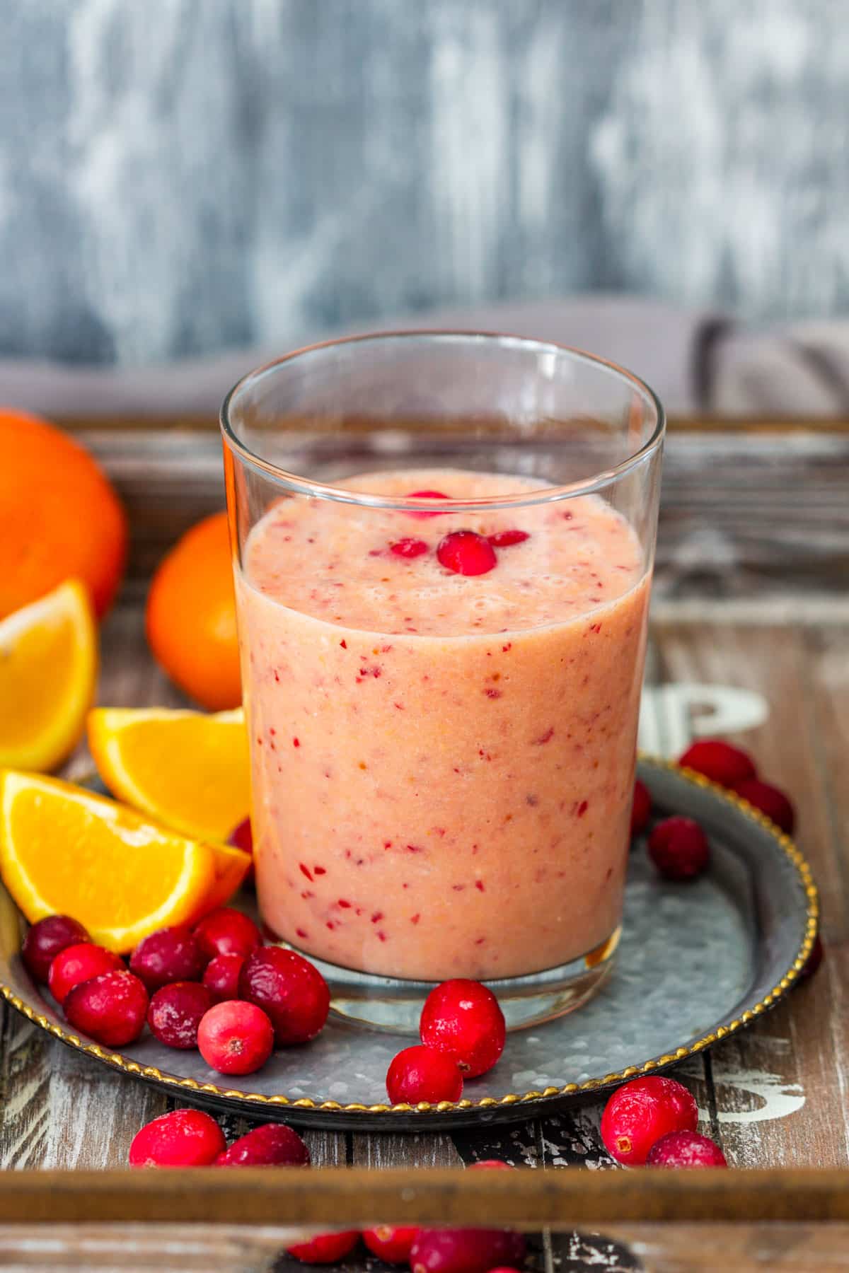 Orange cranberry smoothie in a glass with cranberries and orange wedges on a metal tray.