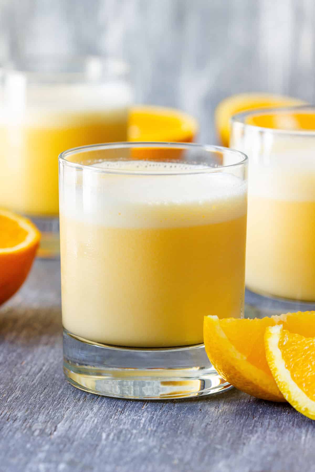 Frothy Orange Julius in a short glass with fresh orange wedges and two more glasses in the background.
