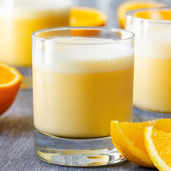 Frothy Orange Julius in a short glass with orange wedges and two blurred glasses behind it.