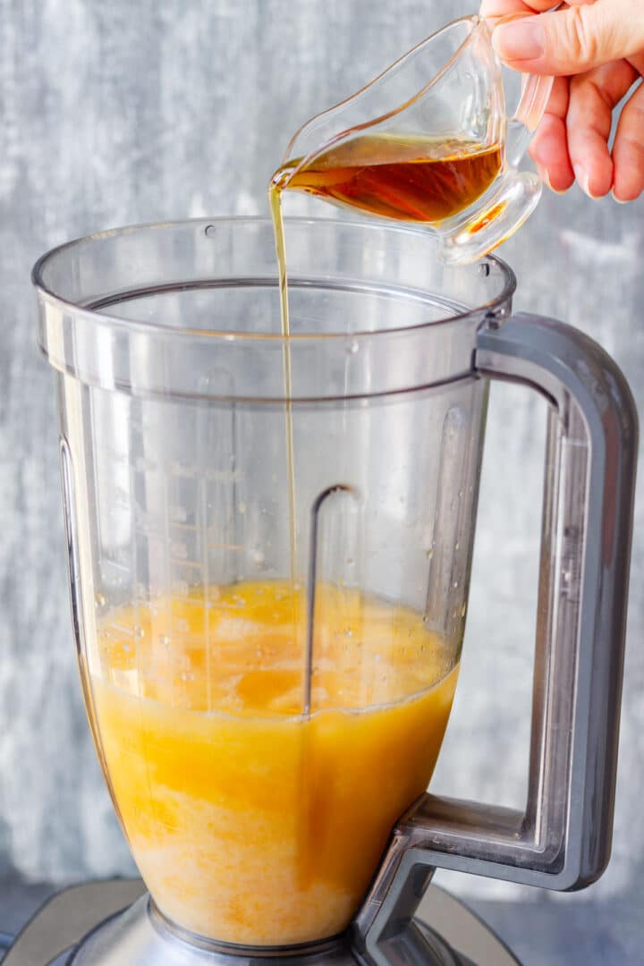 Maple syrup being poured into the orange juice mixture in a blender.