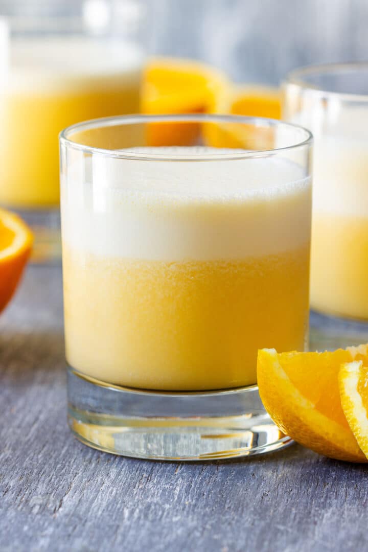 Glass of Orange Julius with a thick foam top and fresh orange wedges.