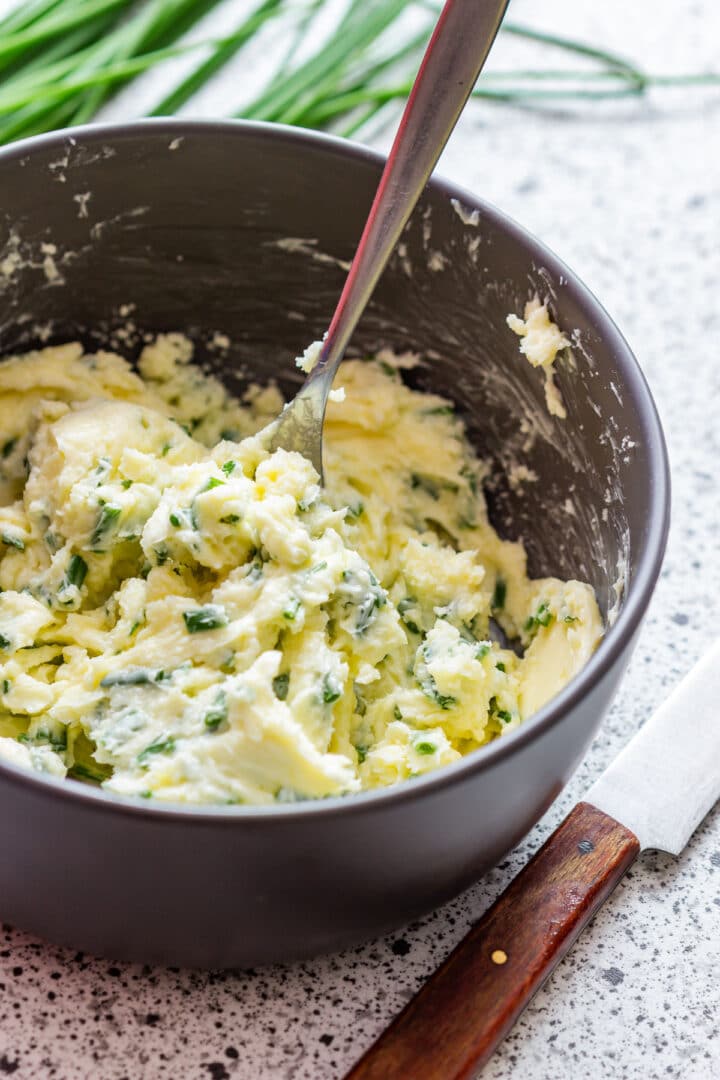 Compound butter flecked with chopped chives in a dark bowl with a fork.