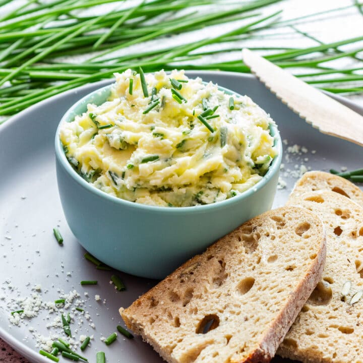 A bowl of Parmesan chive butter with a wooden spreader and slices of seeded bread.