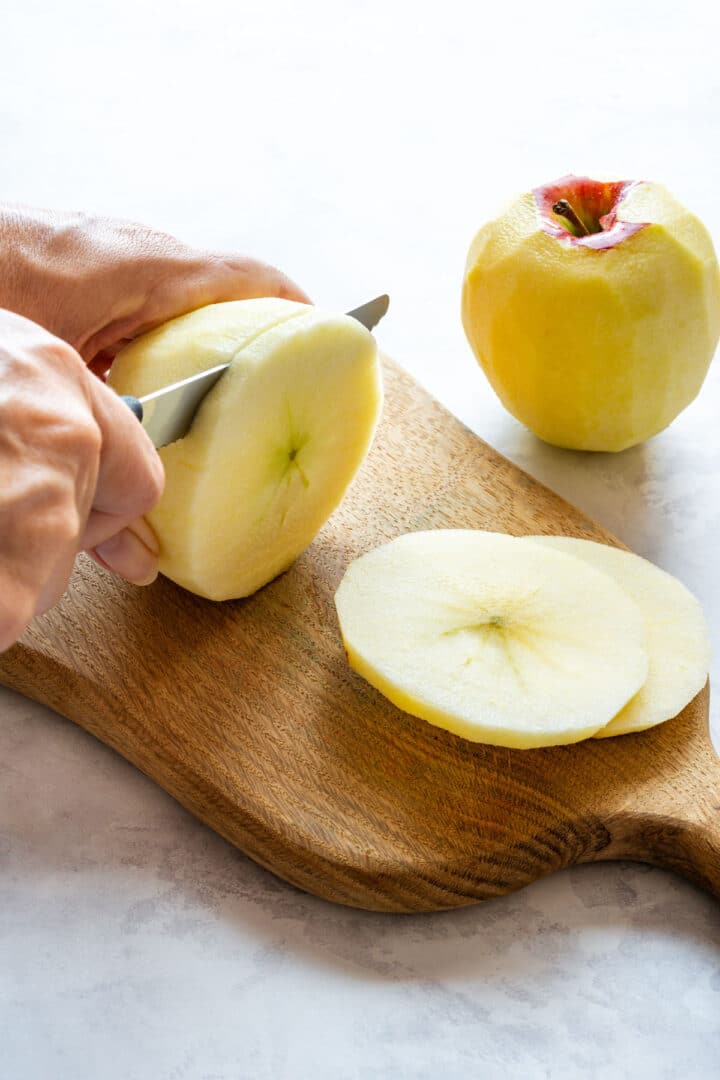 A peeled apple being sliced into thin rounds with a knife, with more apple slices on the cutting board.