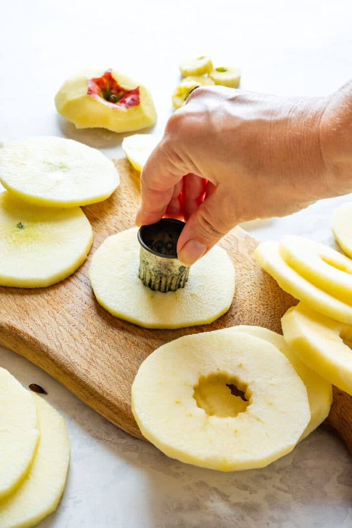 A small round cookie cutter removing the core from a peeled apple slice, with finished apple rings beside it.