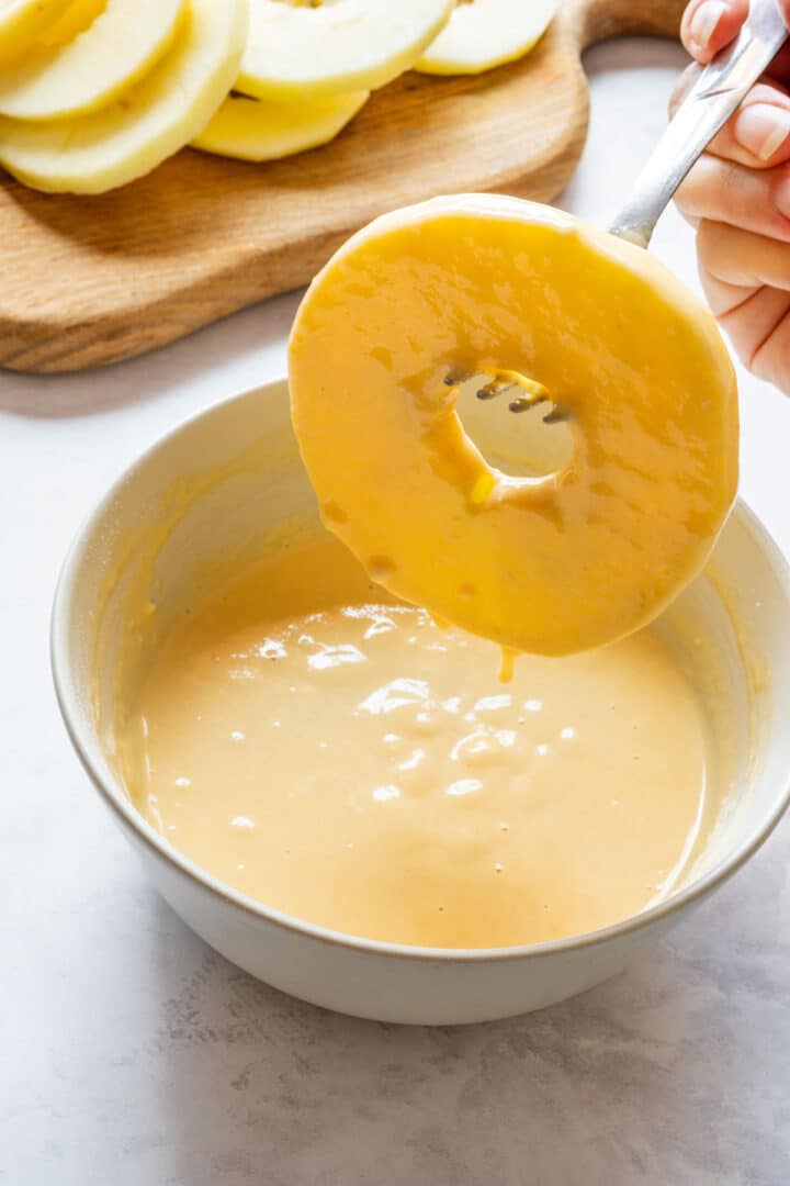 An apple ring lifted on a fork above a bowl of smooth batter, with excess batter dripping back into the bowl.