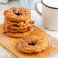 Stack of golden battered apple rings coated in cinnamon sugar, with one ring in front and a mug of milk beside them.