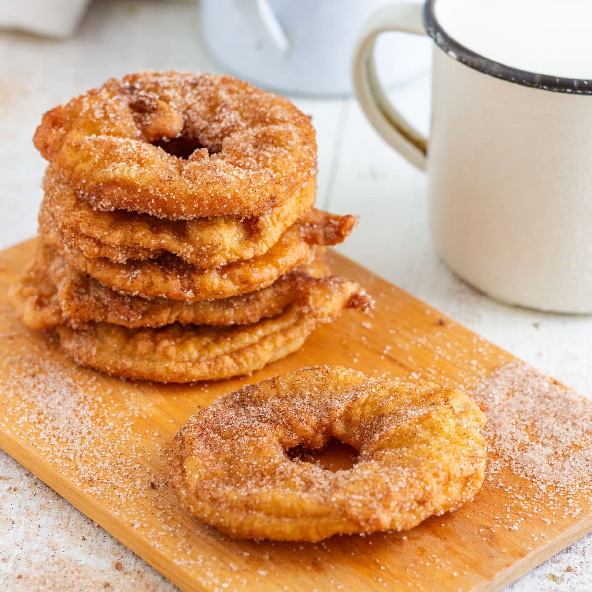 Stack of golden battered apple rings coated in cinnamon sugar, with one ring in front and a mug of milk beside them.