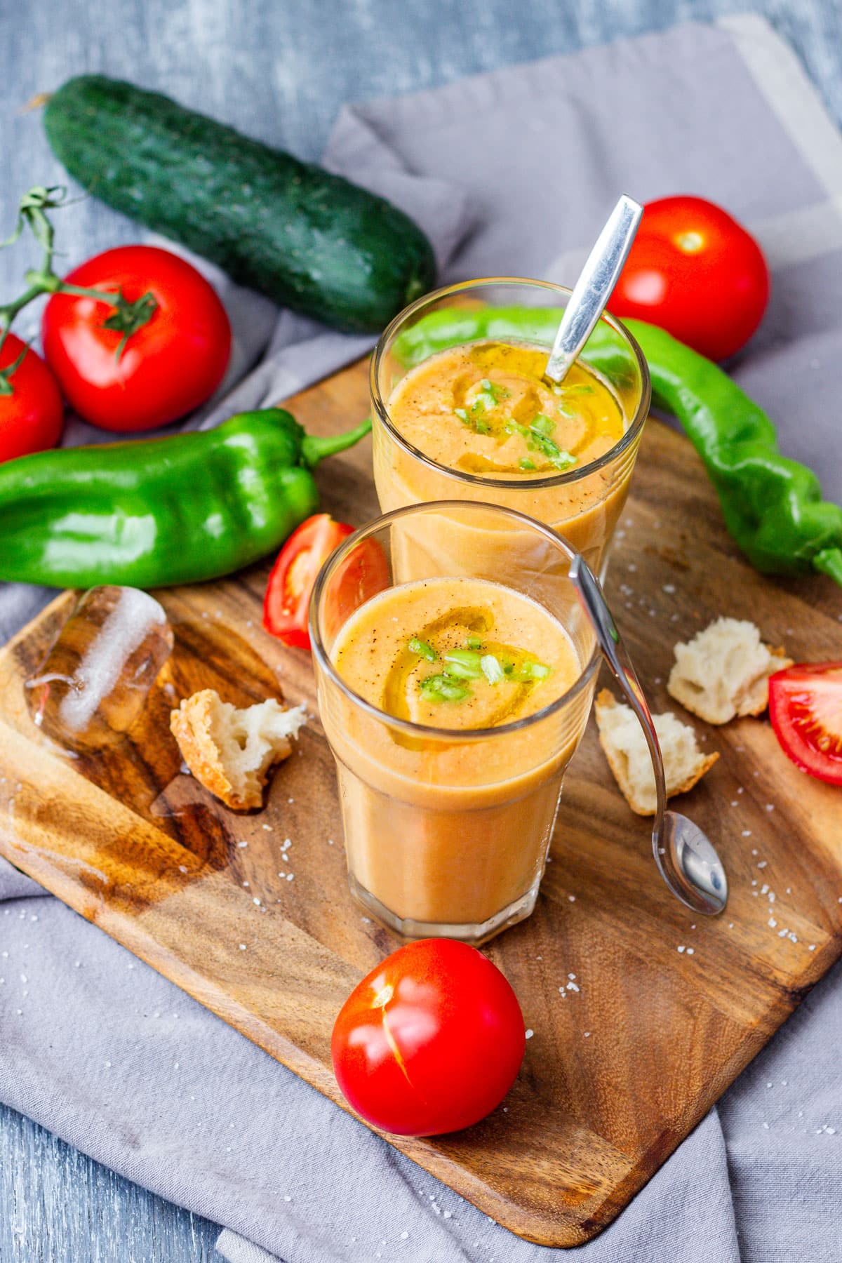 Two glasses of gazpacho soup topped with olive oil and chopped green pepper, on a wooden board with tomatoes, cucumber and bread.