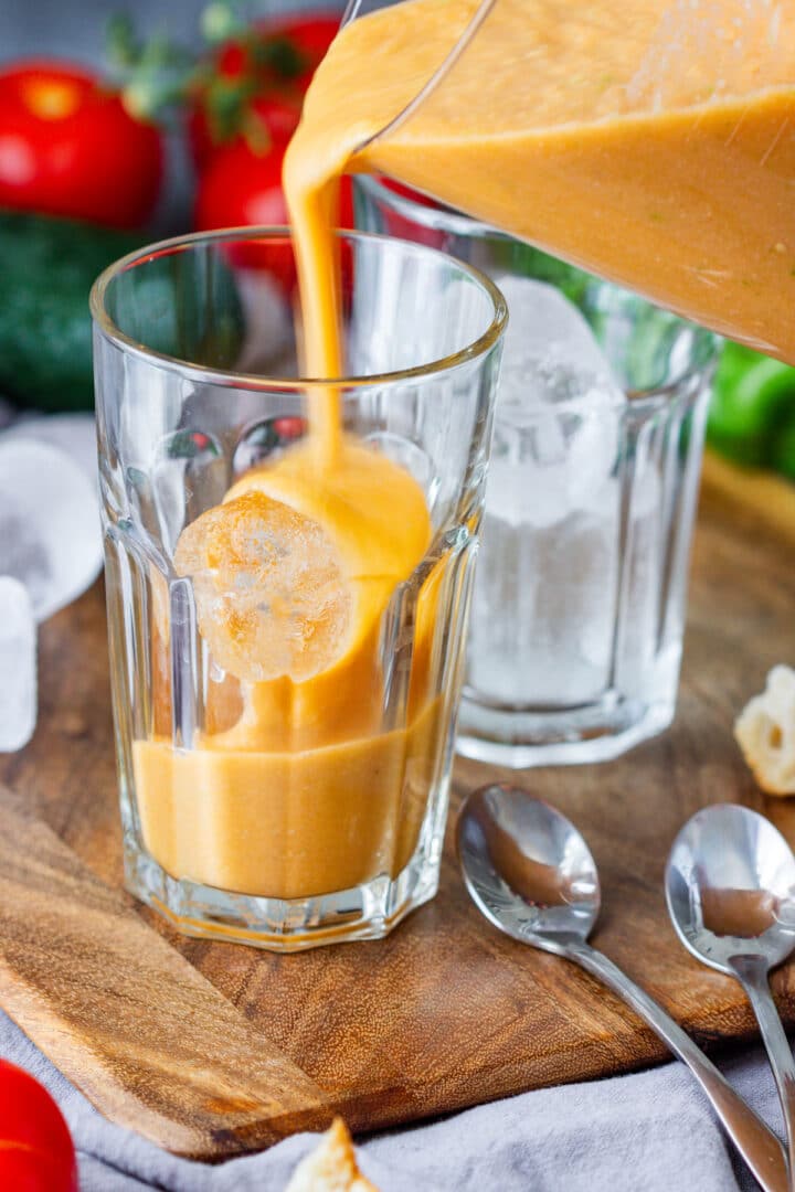 Pouring gazpacho into a glass over a large ice cube, with a second glass and spoons alongside.