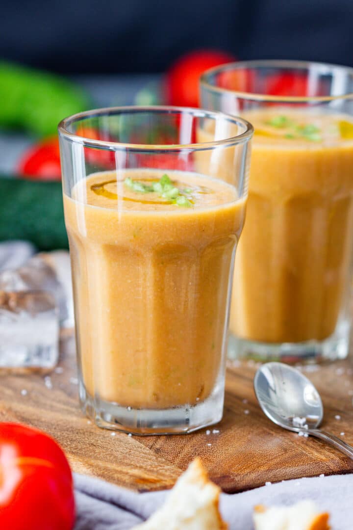 Two glasses of thick cold gazpacho garnished with olive oil and green pepper, with ice and bread nearby.