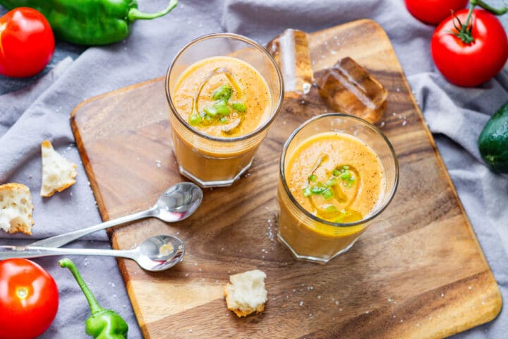 Overhead view of two glasses of gazpacho on a wooden board with spoons, tomatoes and torn bread.