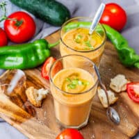 Cold Spanish gazpacho soup served in two glasses with tomatoes, green peppers, cucumber and pieces of bread.