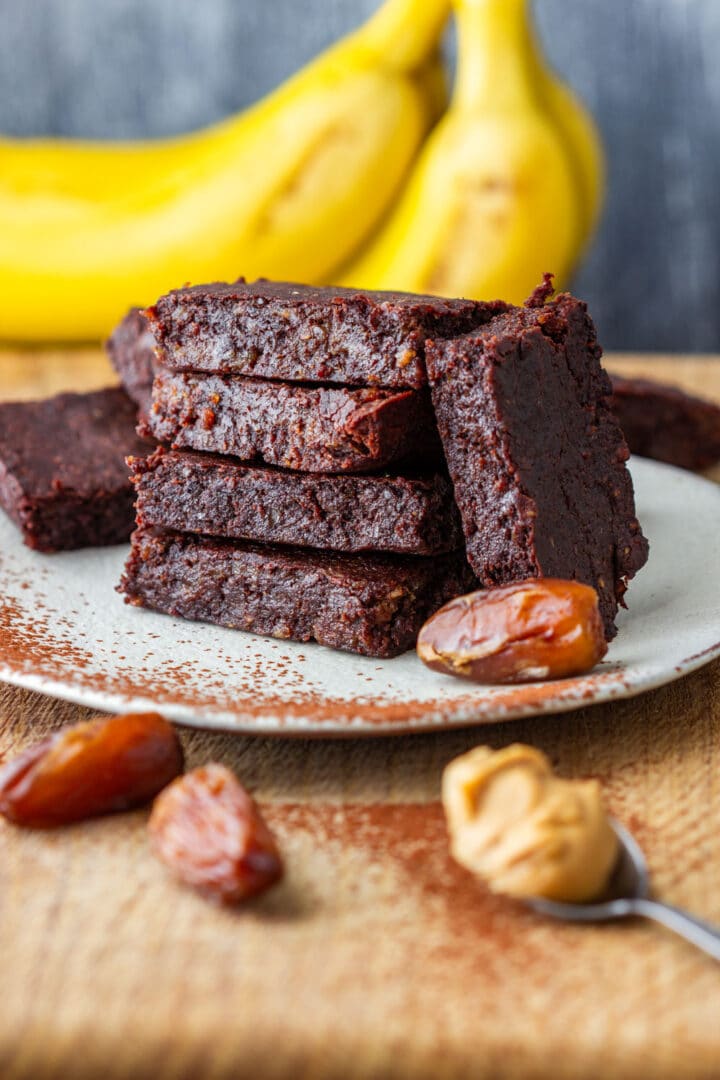 Vegan peanut butter banana brownies made with dates, stacked on a plate with bananas, dates and peanut butter. These flourless, naturally gluten-free brownies are made with cocoa and have a fudgy texture.