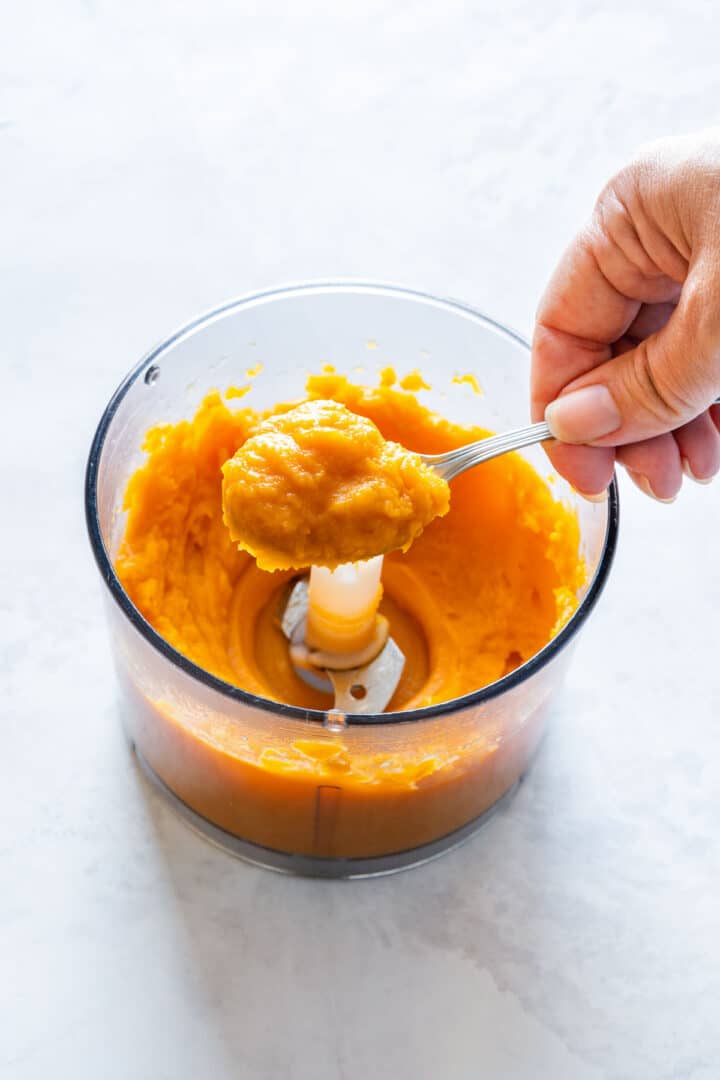 A spoonful of smooth sweet potato purée held above the food processor bowl, showing its thick, creamy texture.