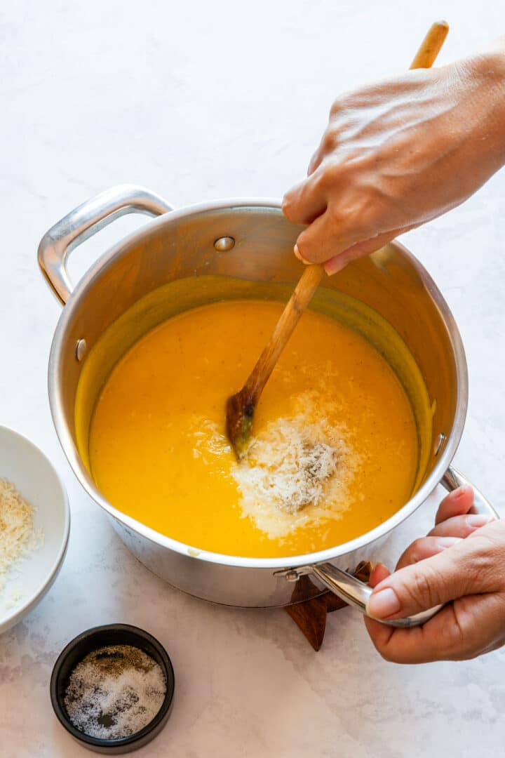Grated Parmesan being stirred into the squash sauce in a pot, with salt and pepper alongside.