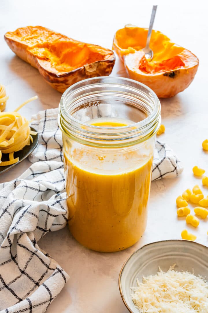 A jar of butternut squash sauce beside roasted squash halves and grated Parmesan.