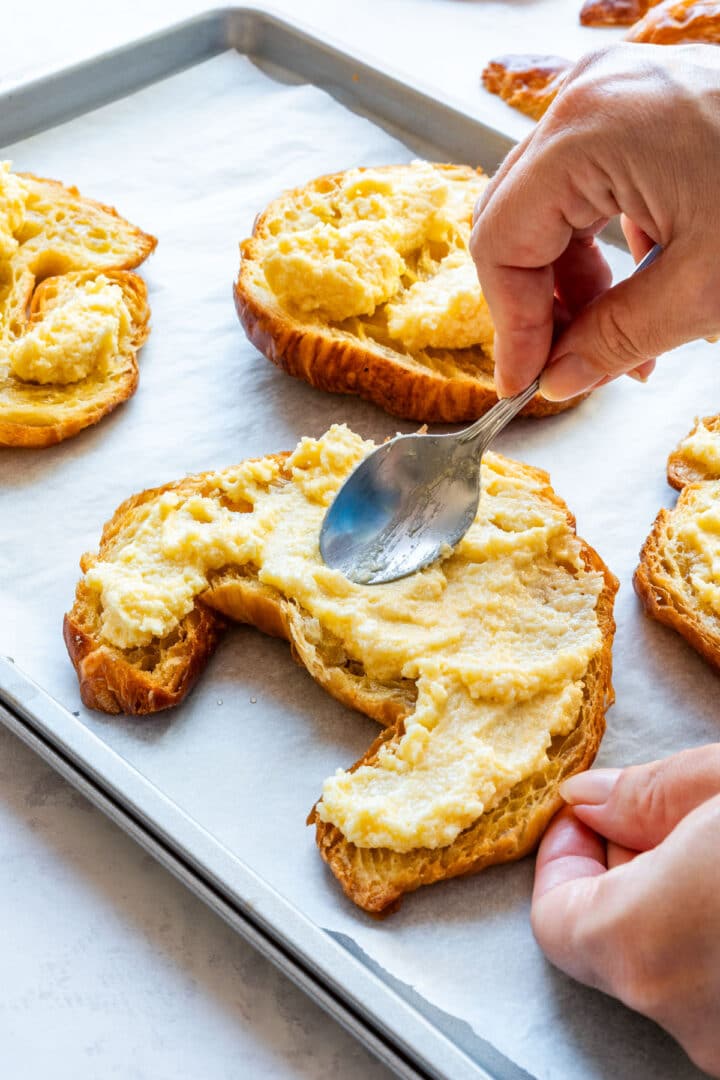 Spreading frangipane over the bottom half of a croissant with a spoon.