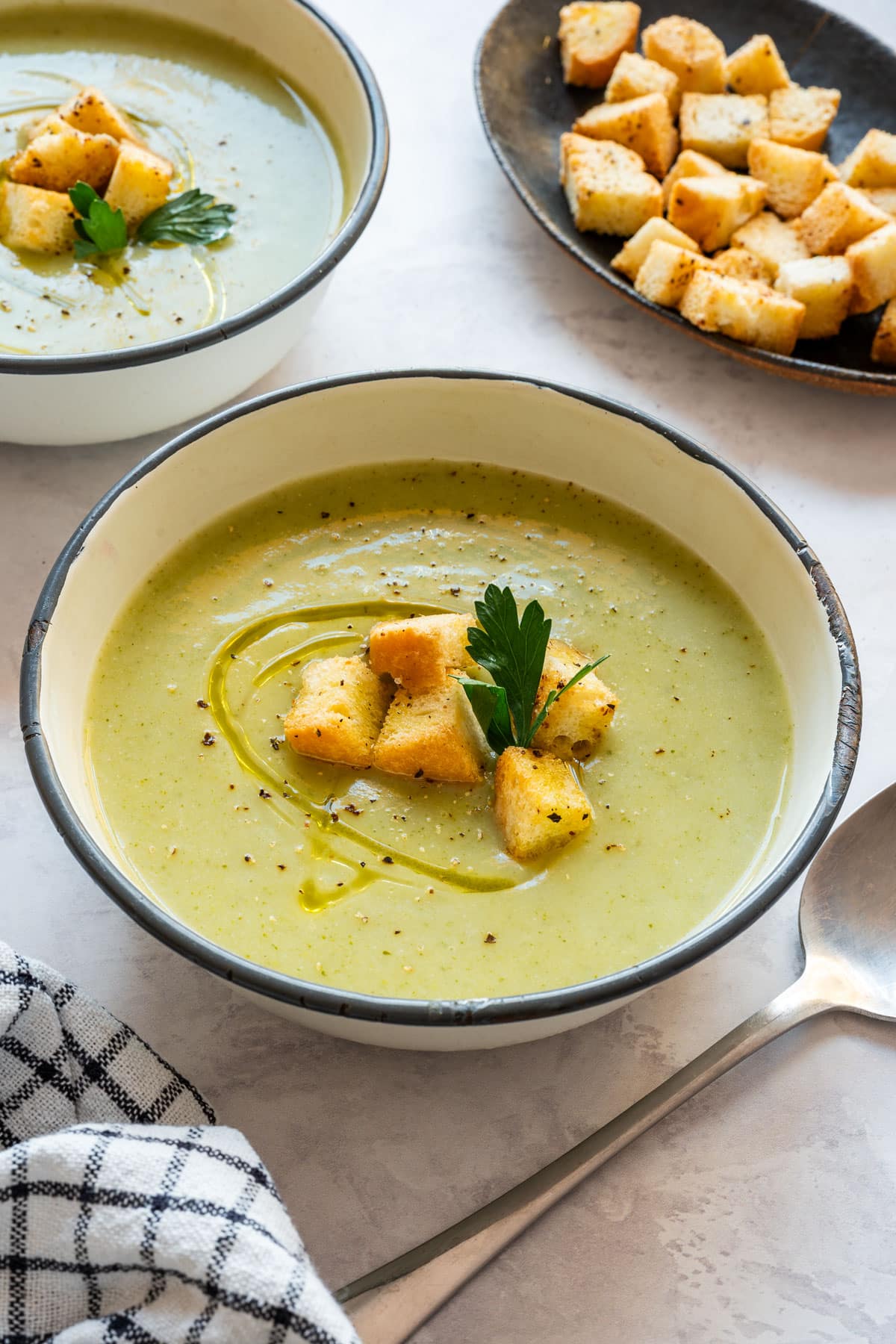 A bowl of broccoli and cauliflower soup topped with golden croutons, fresh parsley and a swirl of olive oil, with another bowl and a plate of croutons behind it.