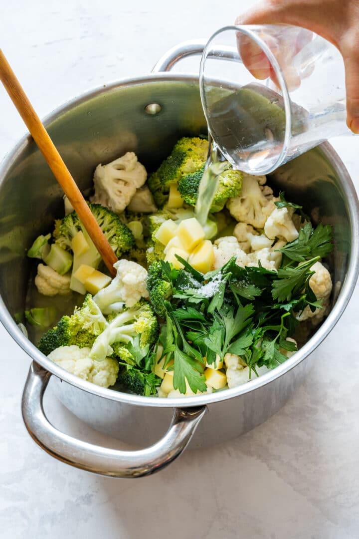 Water being poured over broccoli and cauliflower florets, diced potato and fresh parsley in a large pot.