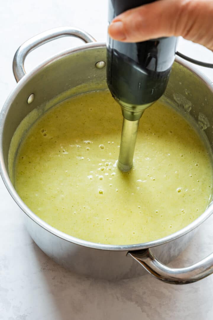 Cooked vegetables being blended into a smooth, pale green soup with an immersion blender.