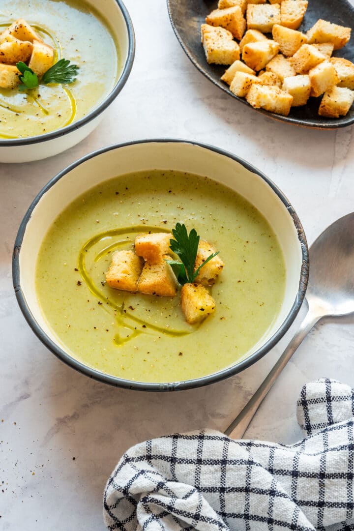 A bowl of smooth soup topped with croutons, with a spoon and a checked kitchen towel beside it.