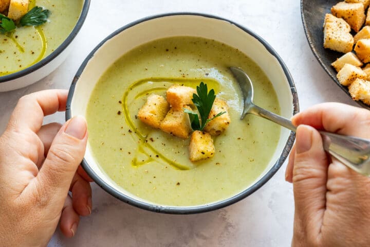 A spoon dipping into a bowl of smooth soup with croutons and parsley on top.